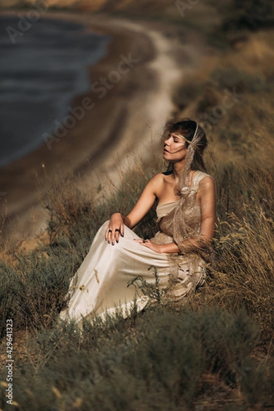 Obraz A beautiful girl in a stylized suit sits on a mountain above the sea, lake on a sunny day.