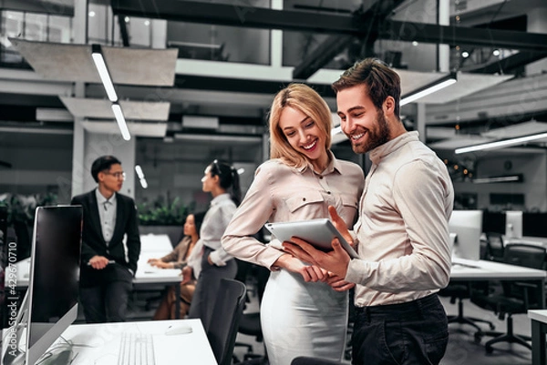 Fototapeta Two handsome business people communicate together and look at the tablet screen standing in a spacious office. The concept of business, finance, projects, teamwork.