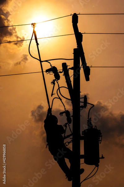 Fototapeta The silhouette of power lineman climbing on an electric pole with a transformer installed. And replacing the damaged hotline clamp, bail clamp, dropout and surge arrester that causes a power failure.