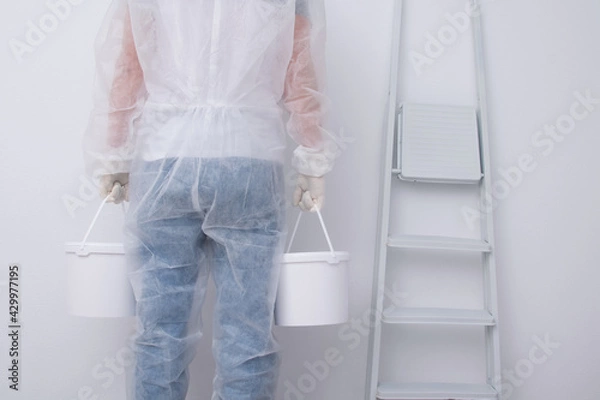 Obraz a man in protective clothing, bringing buckets of paint, against the background of a white wall and a stepladder, rear view