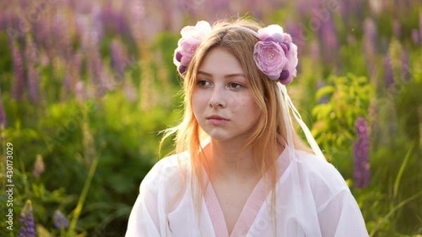 Fototapeta Beautiful blonde girl in a fantasy outfit dress on a field of lupines flowers at sunset in backlight