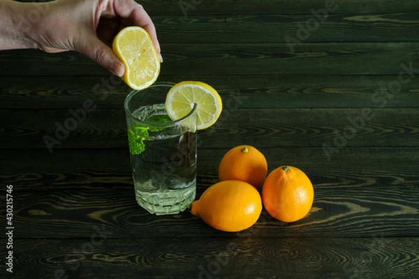 Fototapeta the hand which is squeezing out juice of a half of a lemon both three whole lemons and a segment of a lemon on the edge of transparent glass glass with water and a branch of mint on dark wooden table