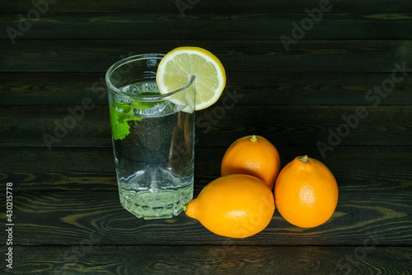 Fototapeta three whole lemons and segment of a lemon on the edge of a transparent glass glass with water and a branch of mint on a dark wooden table from boards