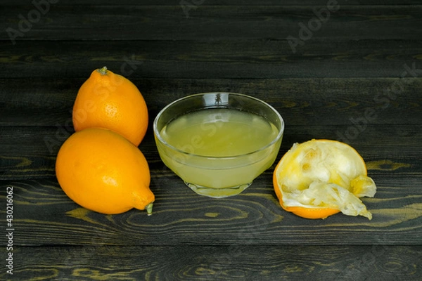 Fototapeta two juicy ripe lemons, a squeezed lemon peel and a glass cup with lemon juice on a dark, coarse wooden background made of boards