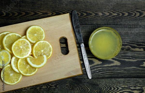 Fototapeta slices of fresh juicy bright yellow lemons laid out on a light wooden cutting board and a glass plate with lemon juice and a knife on a dark wooden table, taken close-up
