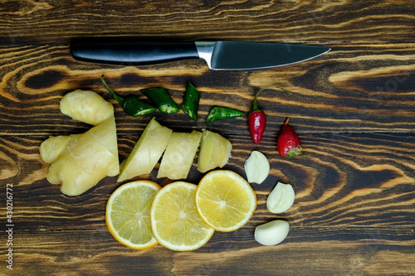 Fototapeta fresh juicy ginger cut into slices, lemon slices, garlic slices, green and red hot peppers and a knife on a dark, coarse wooden background