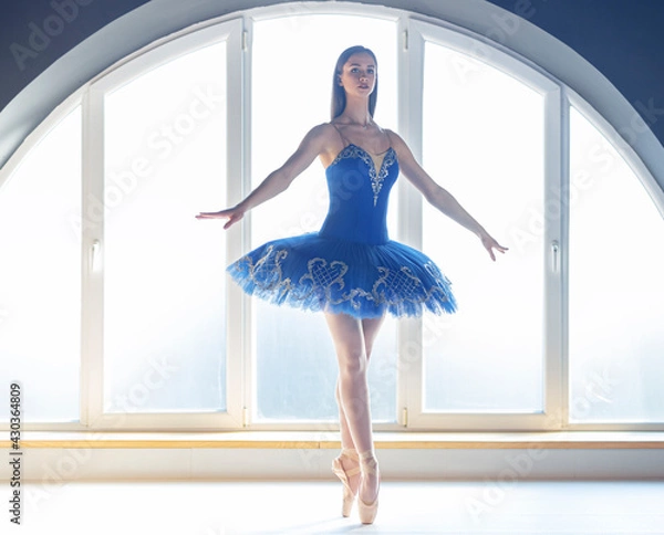 Fototapeta Focused young ballerina dressed in blue tutu costume makes pirouette at ballet studio in front of big round shaped window background in natural ambient daylight atmosphere