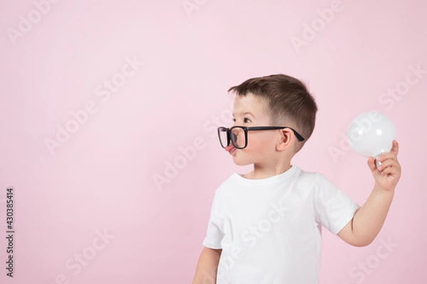 Fototapeta The cute little boy in glasses holds a light bulb and shows his tongue on pink background. Creative thinking concept.