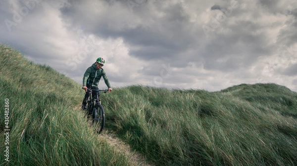 Fototapeta Man with bike in sand dunes with grass at coast in Denmark, dark clouds in dramatic sky.
