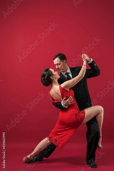 Fototapeta Couple of professional tango dancers in elegant suit and dress pose in a dancing movement on red background. Handsome man and woman dance looking  eye to eye.