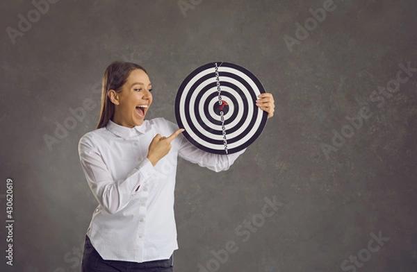 Fototapeta Happy excited smiling young woman holding dartboard target with red dart right in bullseye standing on gray studio background. Business goal, target market, objective, success concept