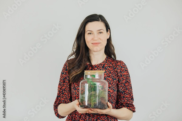 Fototapeta Young woman holds a terrarium in her hands and smiles. Young florist on a white background is holding a large glass container with plants in it. 