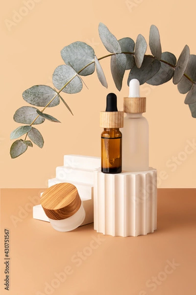 Fototapeta Cosmetic skin care box and two bottles with dropper and wooden covers,from glass staying on the geometrical podiums.Empty space for standing products.Eucalyptus branch above.Pastel beige color.