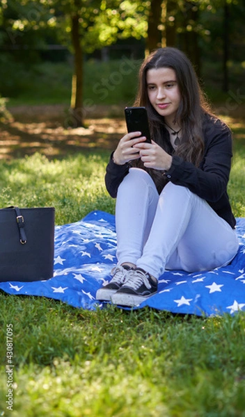 Fototapeta young woman with long hair in a black shirt and white pants sitting on a blanket of stars on the grass in a park with her bag using her phone to take pictures and video conferencing. influencer nature