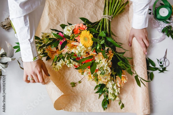 Fototapeta a female florist in a white shirt wraps a bouquet of fresh flowers in paper. Top view