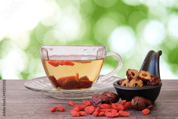 Fototapeta Healthy hot drink brewed from Chinese jujube and Chinese goji berries in glass and sliced Chinese date in the black cup on wooden background with  green blurred garden background. 