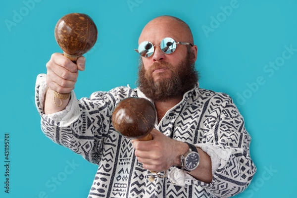 Fototapeta A guy with a bald head in glasses with maracas in his hands. A musician with a full beard and a mustache in a colorful shirt. Man portrait. Coconut Musical Instrument
                               