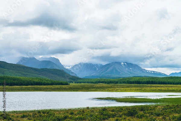 Fototapeta Dramatic landscape with mountain lake and forest on hills in sunlight and snowy mountains in low clouds in changeable weather. High mountains in cloudy sky and lake near green grasses and forest hills