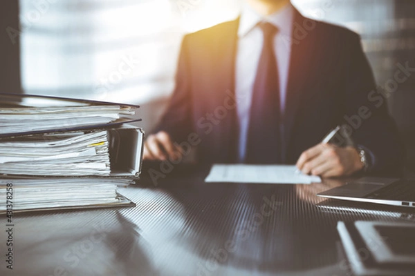 Fototapeta Binders with papers are waiting to be processed by unknown man accountant in blue blazer staying at home during covid pandemic. Taxes and audit concept