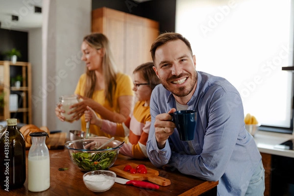 Fototapeta Father is drinking a coffee, smiling and looking at camera while his little daughter is cooking