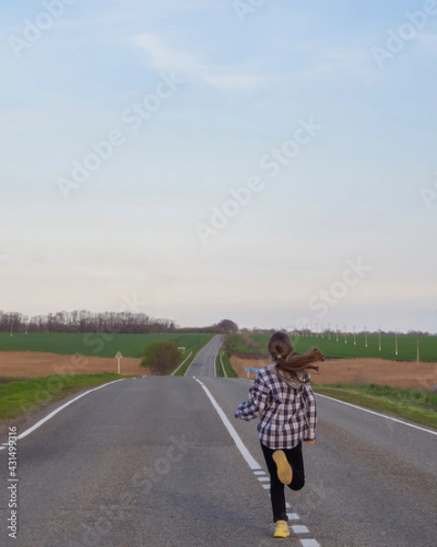 Obraz A young girl runs along the road. A girl with long blond hair runs away alone on a deserted road that goes into the distance among the fields. Healthy lifestyle.