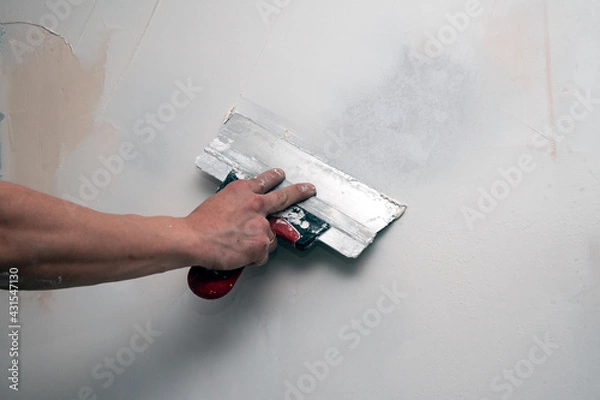 Fototapeta The hand of the builder puttying the wall with putty, leveling the wall with a spatula