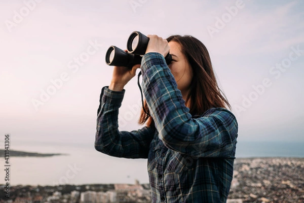 Obraz Portrait of a young female tourist looking through binoculars at sunset.