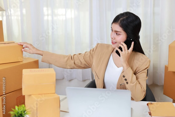 Fototapeta Young asian smart and happy business woman in casual dress working from home with laptop computer and smartphone with online purchase shopping order and box packaging on table (new normal concept)