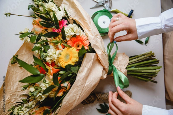 Fototapeta a female florist ties a green ribbon bow on a bouquet of flowers wrapped in craft paper on the desktop. Top view.