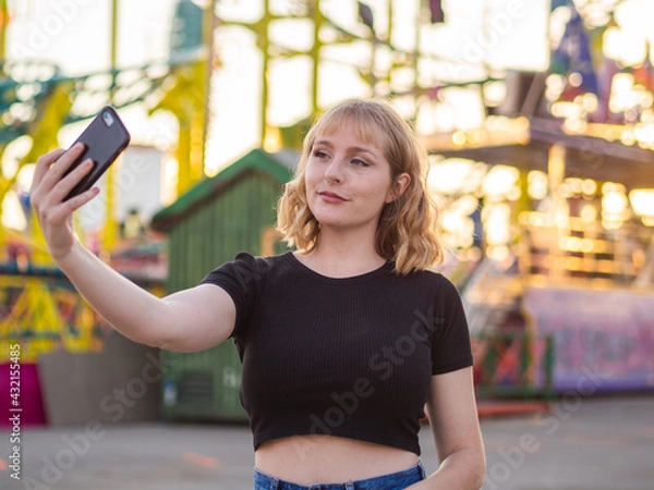 Fototapeta Mujer joven haciéndose un selfie en una feria o parque de atracciones durante la puesta de sol 