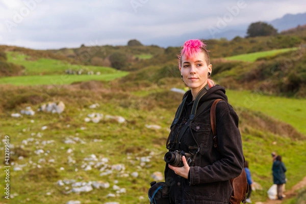 Obraz Young woman with pink hair, punk aesthetics and androgynous features holding a camera in the middle of a natural environment. Alternative and rebellious style traveler.