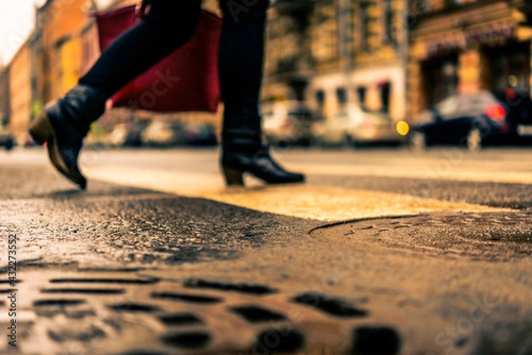 Fototapeta Rainy day in the big city, the woman with a red bag crosses the road on a pedestrian crossing. Close up view of a hatches at the level of the asphalt