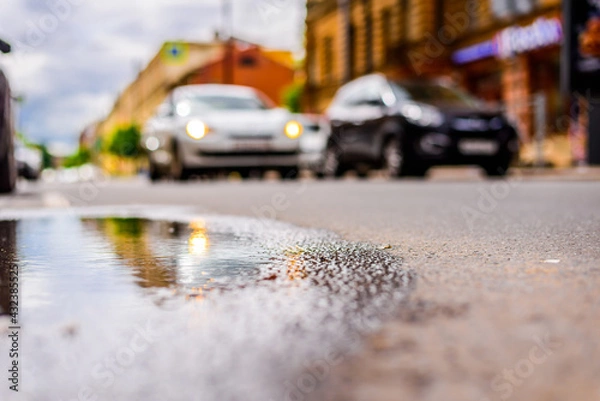 Fototapeta Sunny day after rain in the city, parked cars near the store and a passing car. Close up view from the level of the puddle on the pavement