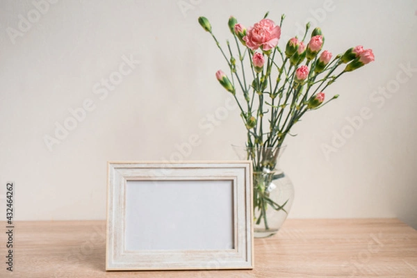 Fototapeta Portrait white picture frame mockup on wooden table. Modern ceramic vase with carnation. White wall background. Scandinavian interior. 