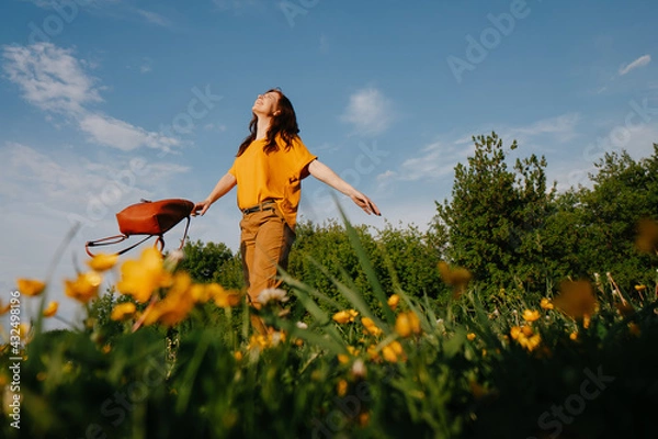 Fototapeta A young dark-haired woman in a yellow dress, with open arms, walks across the field looking at the blue, sunny sky. Wildflowers, summer time.