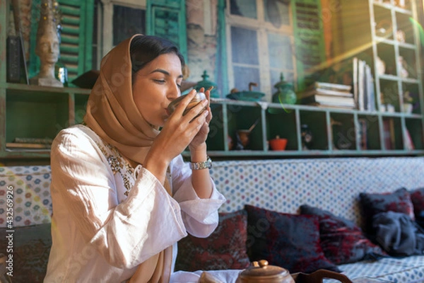Fototapeta Young smiling muslim woman with hijab is sitting in an oriental tea bar and drinking a hot beverage.