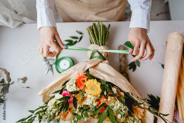 Fototapeta a female florist ties a green ribbon bow on a bouquet of flowers wrapped in craft paper on the desktop. Top view.