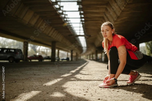 Fototapeta Side view of fit young woman tying shoe laces while standing under the bridge, ready for running.