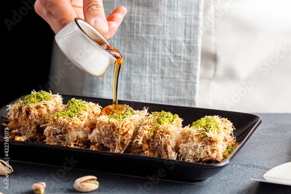 Fototapeta A caucasian chef is pouring thick sweet syrup from a porcelain pitcher onto slices of Turkish kadayif dessert. This is made using shredded phyllo dough walnuts and pistachio.