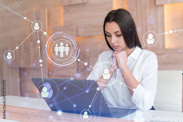 Fototapeta Attractive businesswoman in white shirt at workplace working with laptop to hire new employees for international business consulting. HR, social media hologram icons over office background