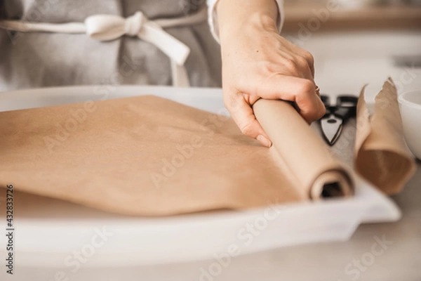 Fototapeta The girl unfolds parchment paper for baking on a light baking sheet. Cooking at home, handmade sweets