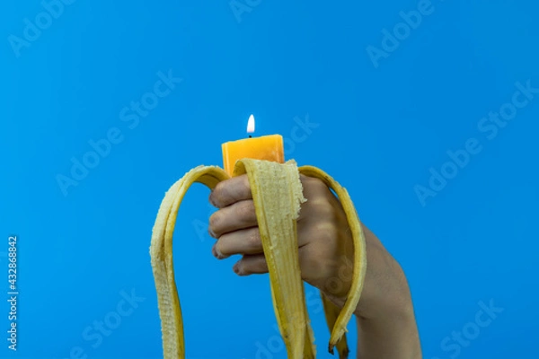 Fototapeta Mano di giovane ragazza tiene in pugno una banana sbucciata con all'interno una candela accesa dal basso su sfondo blu