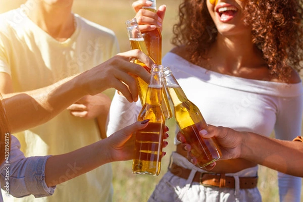 Fototapeta Group having fun enjoying refreshing beverage. Young men and women drink beer on festival, birthday . Happy friends spending free time together in park sitting on grass, drinking beer and chatting.