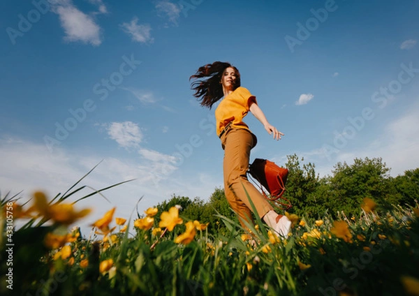 Fototapeta A young slender girl with curly hair developing in the wind runs in a meadow with yellow field dandelions in the park. Allergy free concept.