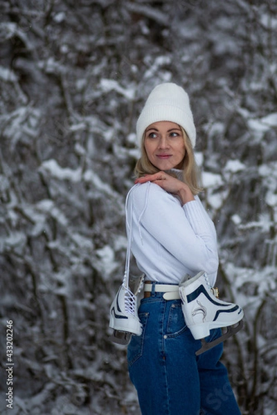 Fototapeta Young blond beautiful female with white ice skates in her hand in winter snowy forest.