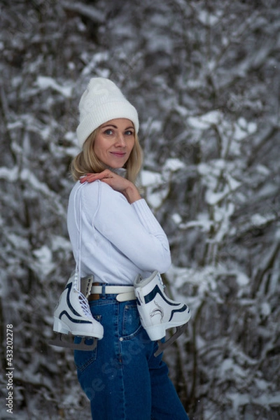 Fototapeta Young blond beautiful female with white ice skates in her hand in winter snowy forest.