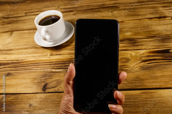 Fototapeta Hand with modern smartphone and cup of coffee on a rustic wooden table