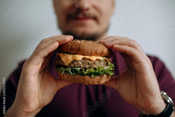 Fototapeta a burger in the hands of a bearded young man. a hungry guy holds a cheeseburger before eating it. a happy man holds a burger in front of him with both hands. copy space