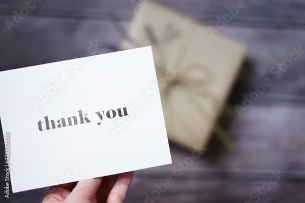 Fototapeta Woman's hand holding a thank you card with a present on wooden background. Special thank you note. Surprise gift. 