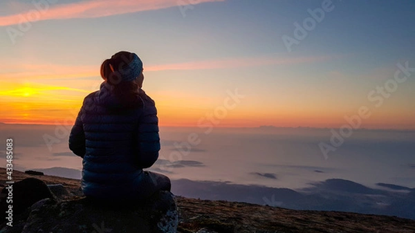 Fototapeta A woman with a headband sitting on top of Babia Gora, Poland, and enjoying the panoramic view on sun rising above the horizon. There are thick clouds below. The sky is pink and orange. Calmness
